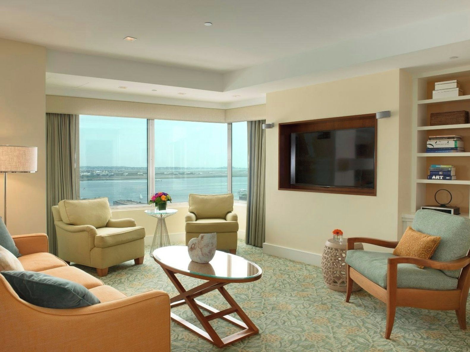 A hotel room with a large window offering a view of the water, two armchairs, a sofa, a coffee table, and a television.