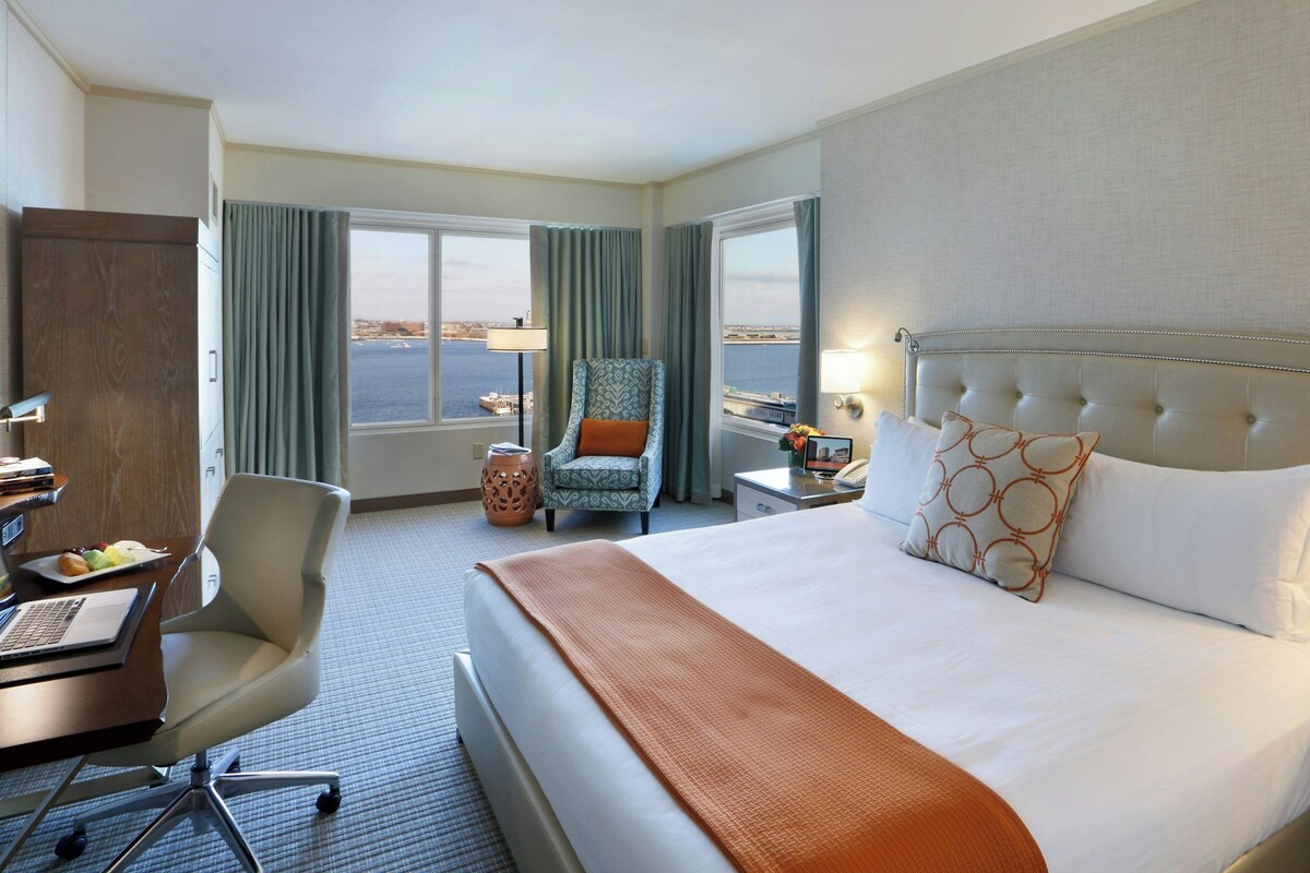 A hotel room with a bed, a desk with a laptop, a chair, a nightstand, and a view of the city through the window.