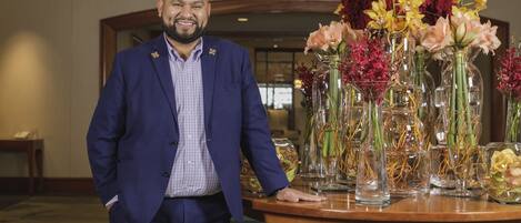 A man in a blue suit standing next to a round wooden table with floral arrangements.