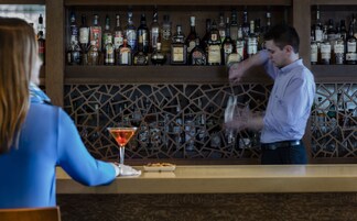 A bar area with a bartender preparing a drink. There are shelves with various liquor bottles behind the bartender.