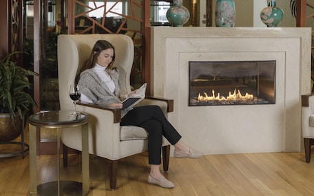 A woman seated in a chair reading a book in a room with a fireplace, wooden floor, and decorative wall panels.