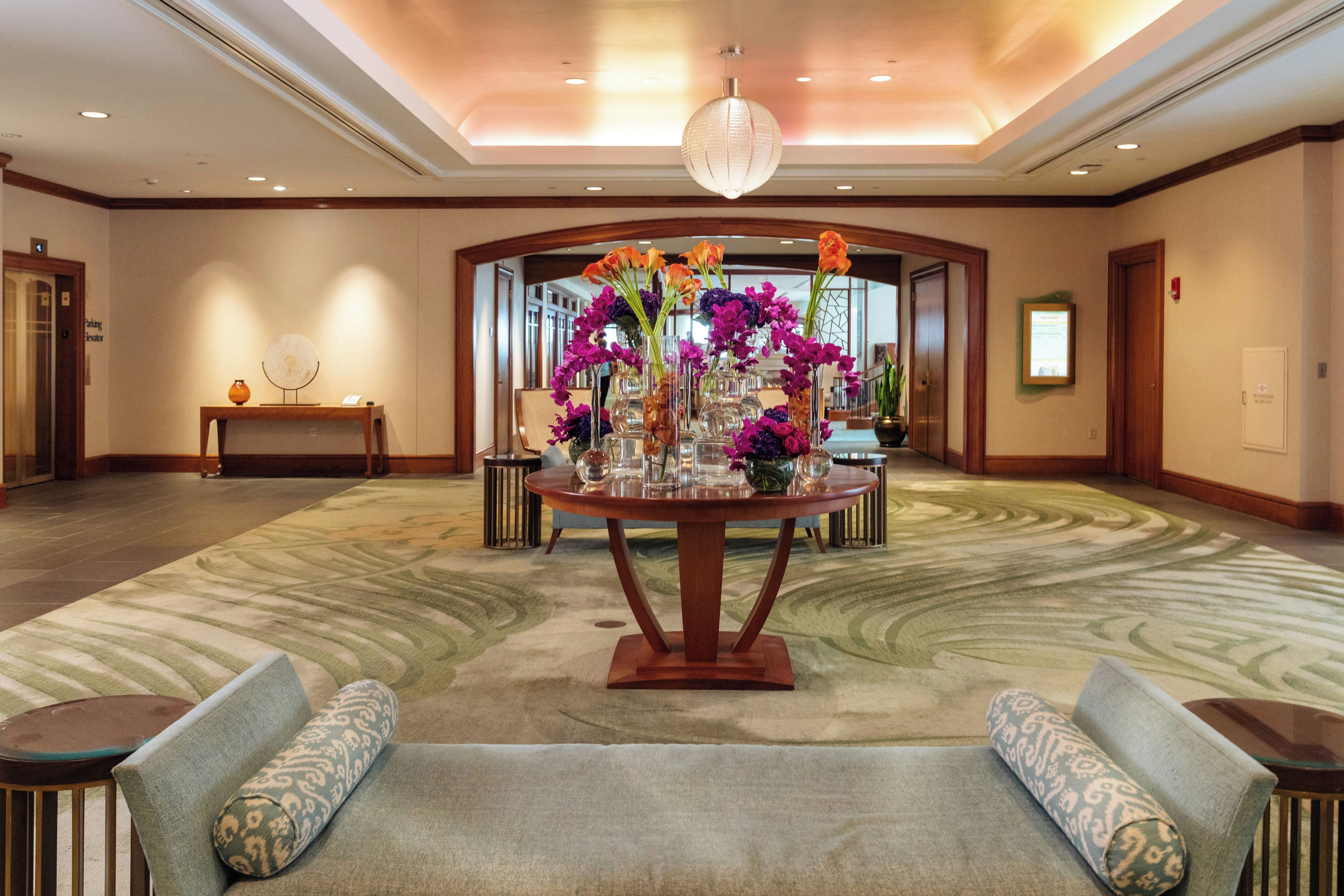 A spacious lobby with a central round table adorned with floral arrangements, flanked by two sofas and a small table with a vase.