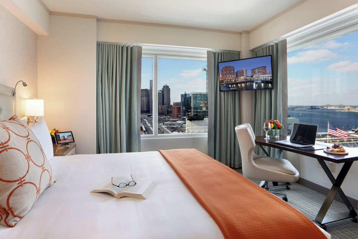 A hotel room with a bed, a desk with a laptop, a TV, and a view of a cityscape.