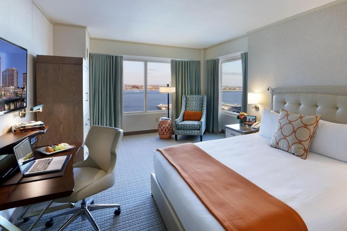 A hotel room with a bed, desk, chair, and a view of the cityscape through the window.
