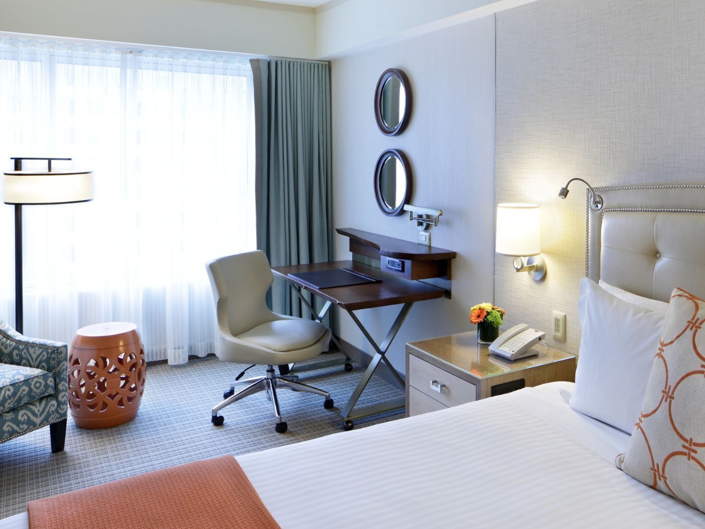Una habitación de hotel con una cama, un escritorio, una silla, una lámpara, un teléfono y una mesita de noche con un florero.