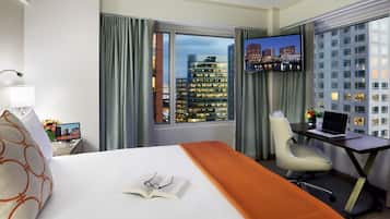 A hotel room with a bed, a desk, a chair, a TV, and a view of a cityscape at night.
