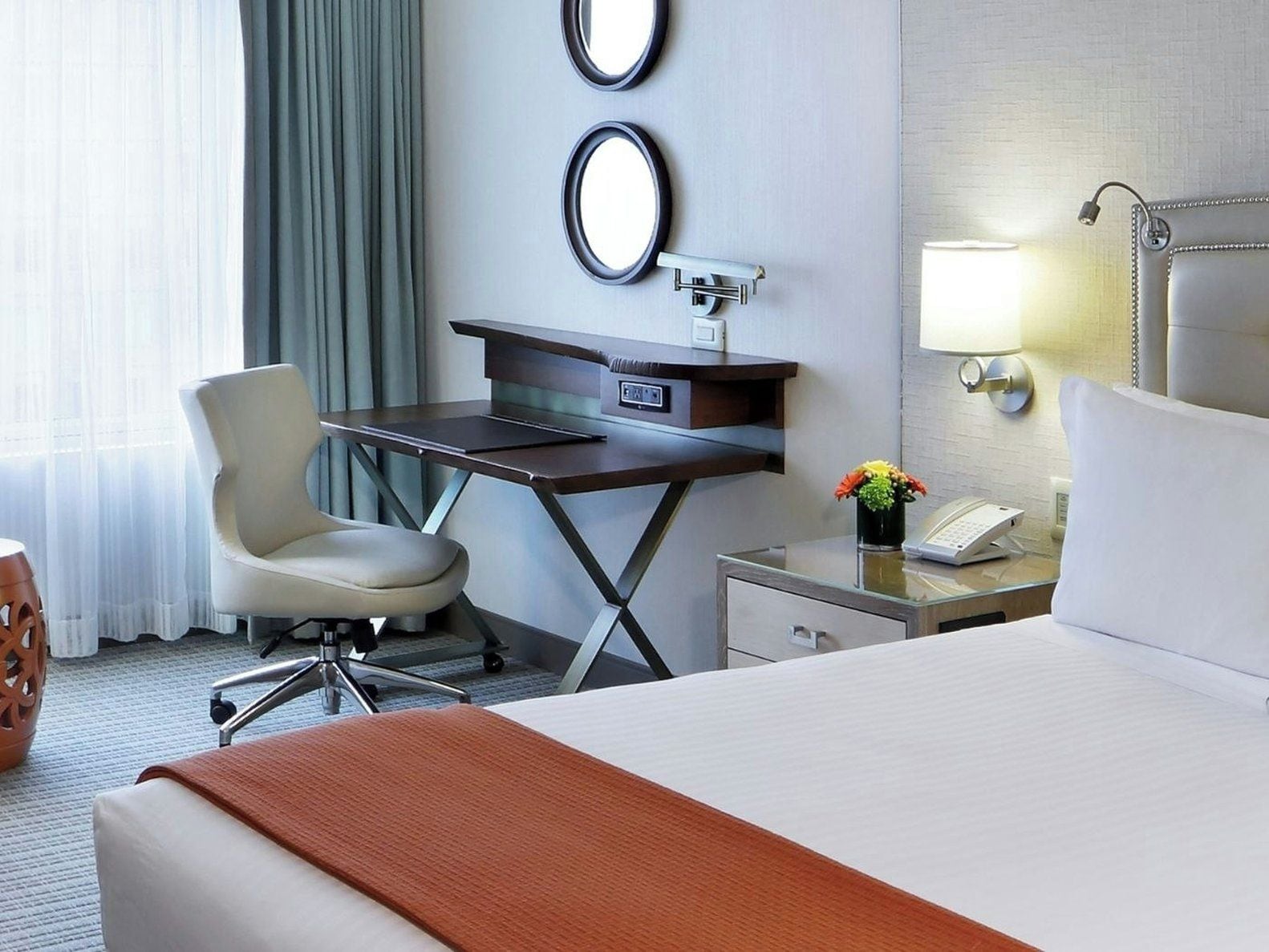 A hotel room with a bed, desk, chair, and a small table with a lamp and phone.