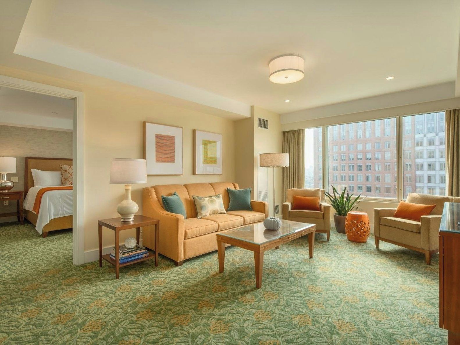 A hotel room with a bed, a sofa, a coffee table, a side table, and a large window with a city view.