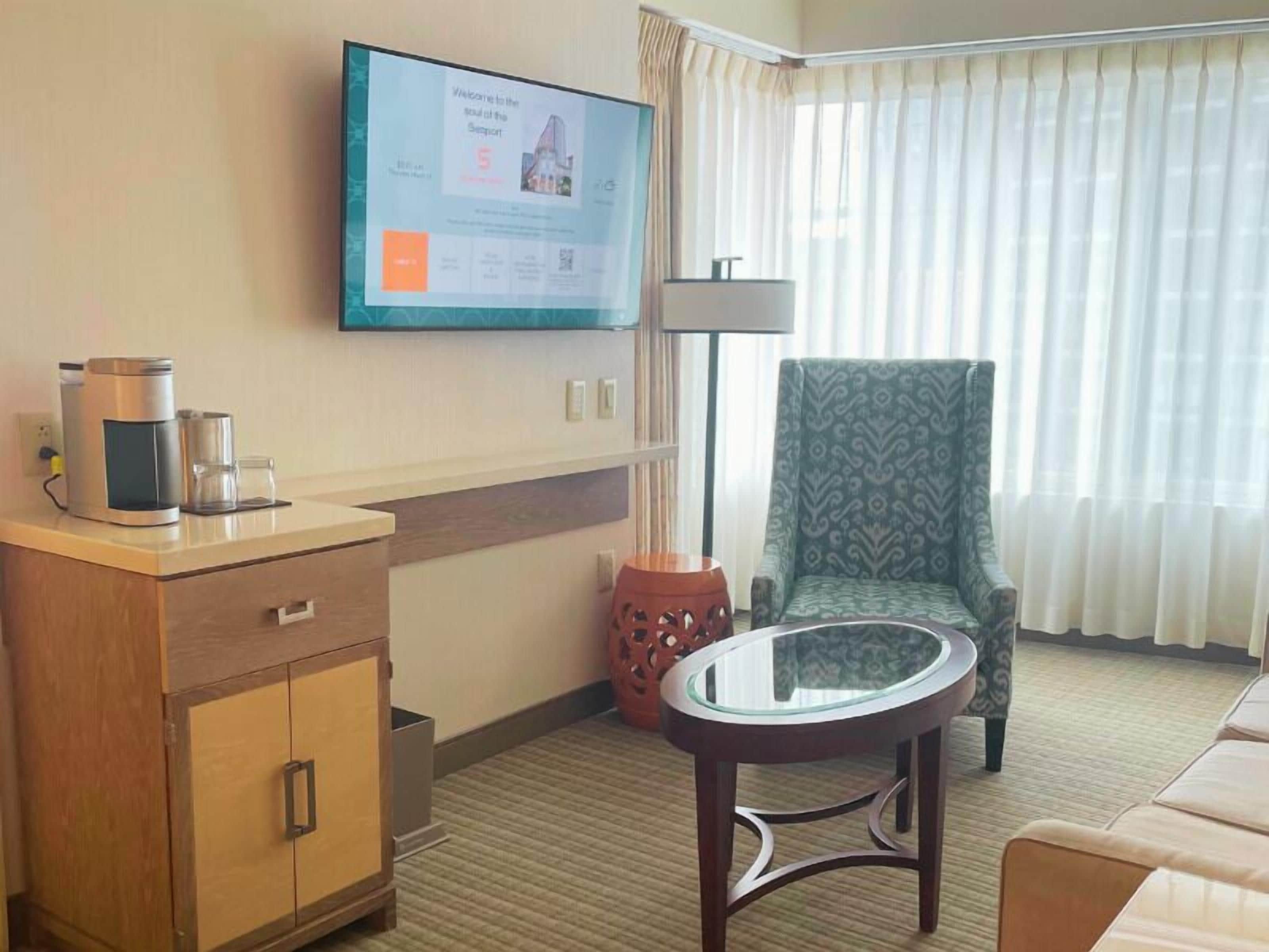 A hotel room with a wooden cabinet, a glass table, a patterned armchair, and a television displaying a welcome message.