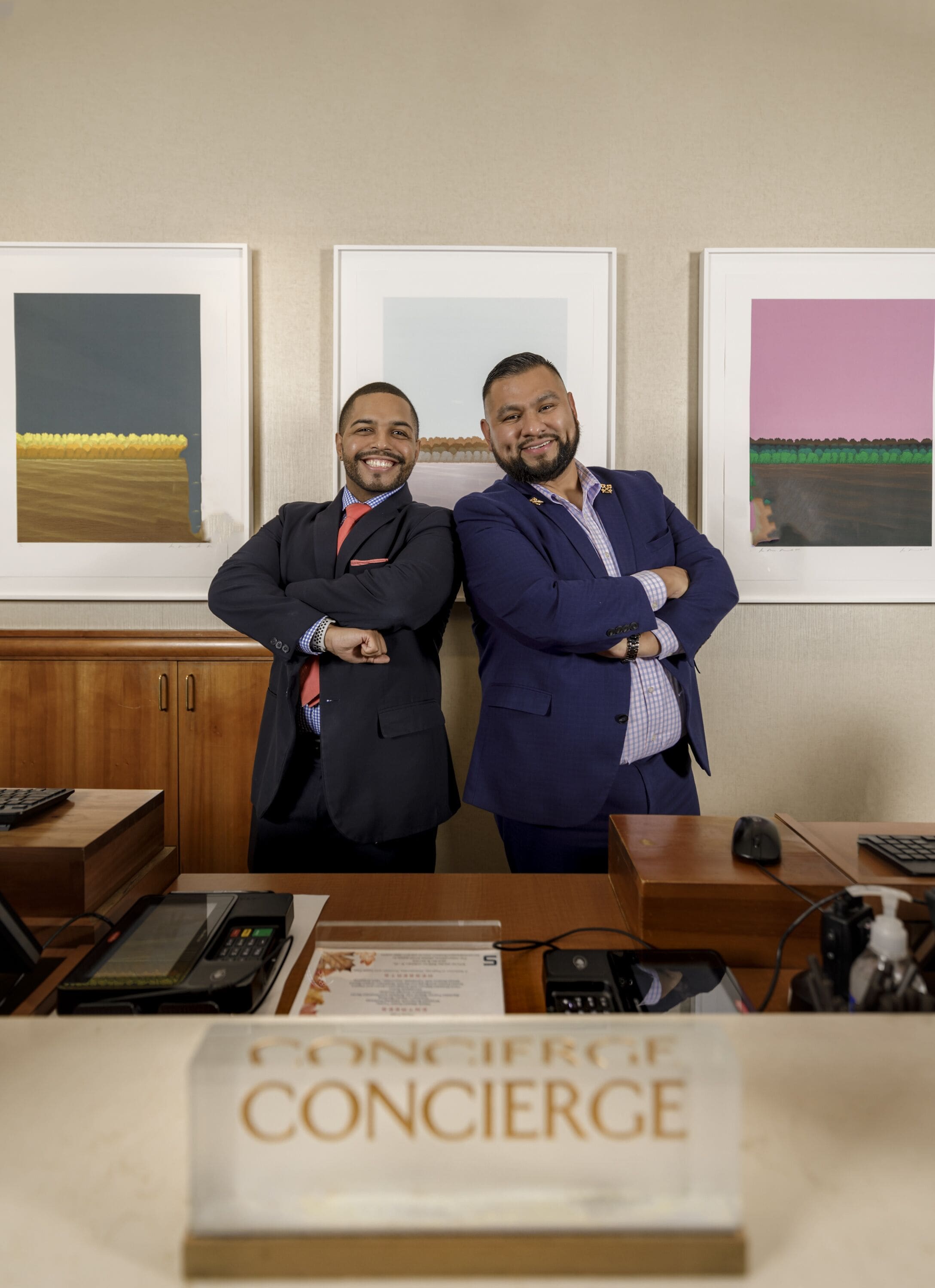 Deux hommes en costume, debout dans un bureau. Des œuvres d’art encadrées ornent le mur, et une plaque sur le bureau indique « CONCIERGE ».