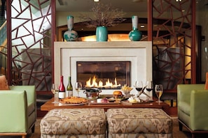 Una chimenea con fuego, rodeada por una mesa de comedor con comida y bebidas, y dos sillones verdes.