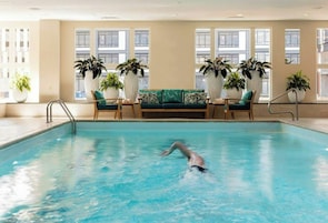 An indoor swimming pool with a person swimming, surrounded by large windows and potted plants.