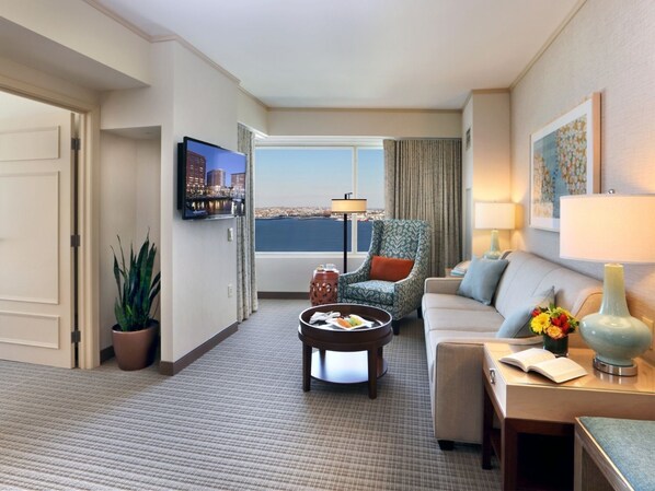 A hotel room with a large window offering a city view, a television, a patterned armchair, a sofa, and a small table with a vase of flowers.