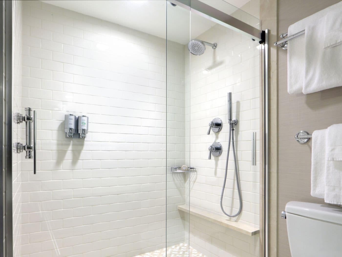 Un área de ducha con paredes de azulejos blancos y puerta de cristal, con un inodoro blanco visible a través del cristal.