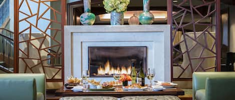 A fireplace with decorative vases, a dining table with food and drinks, and patterned ottomans.