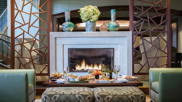Una chimenea con jarrones decorativos, una mesa de comedor con comida y bebidas, y otomanas estampadas.
