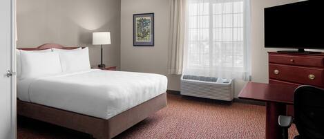 Una habitación de hotel con cama, escritorio, silla, televisor, ventana con cortinas, aire acondicionado instalado en la pared y un cuadro enmarcado.