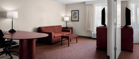 Quarto de hotel com carpete vermelho, sofá, mesa pequena, escrivaninha com cadeira, luminária, quadro emoldurado na parede e televisão sobre um suporte.