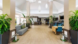 A spacious lobby with a high ceiling, modern pendant lighting, a central seating area, and a kitchen in the background. - Hilton Garden Inn Lake Mary (Lake Mary)