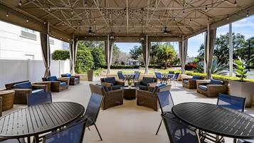 An outdoor patio area with wicker furniture, round tables, and chairs, featuring a view of a landscaped garden and a building in the background.