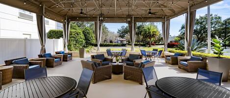 An outdoor patio area with wicker furniture, round tables, and chairs, featuring a view of a landscaped garden and a building in the background.