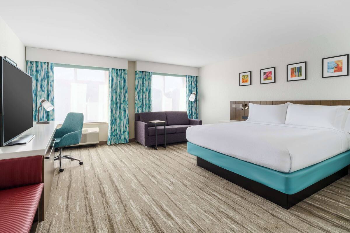 A hotel room with a bed, a desk with a computer, a chair, a TV, and a window with curtains.