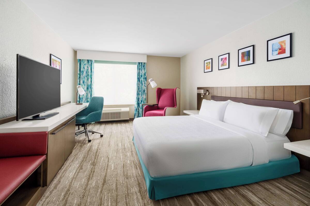 A hotel room with a bed, a desk with a TV, a chair, a window with curtains, and framed pictures on the wall.