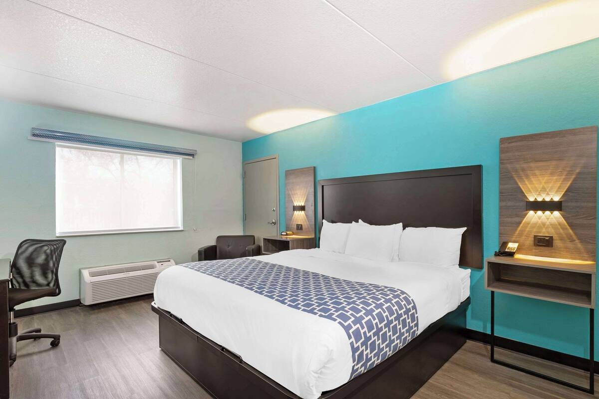 Habitación de hotel con una cama grande, un escritorio, una silla, un ventanal y una lámpara en la pared.