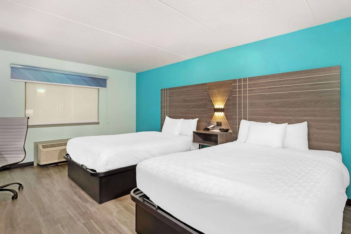 Habitación de hotel con dos camas, una mesita de noche con lámpara, una ventana con persianas y un calentador fijado a la pared.