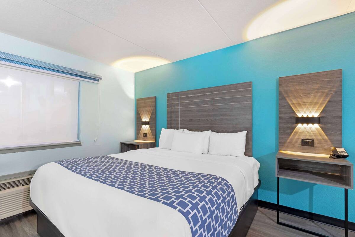 Habitación de hotel con una cama grande, dos mesitas de noche con lámparas, un ventanal con persianas y una pared de acento azul.