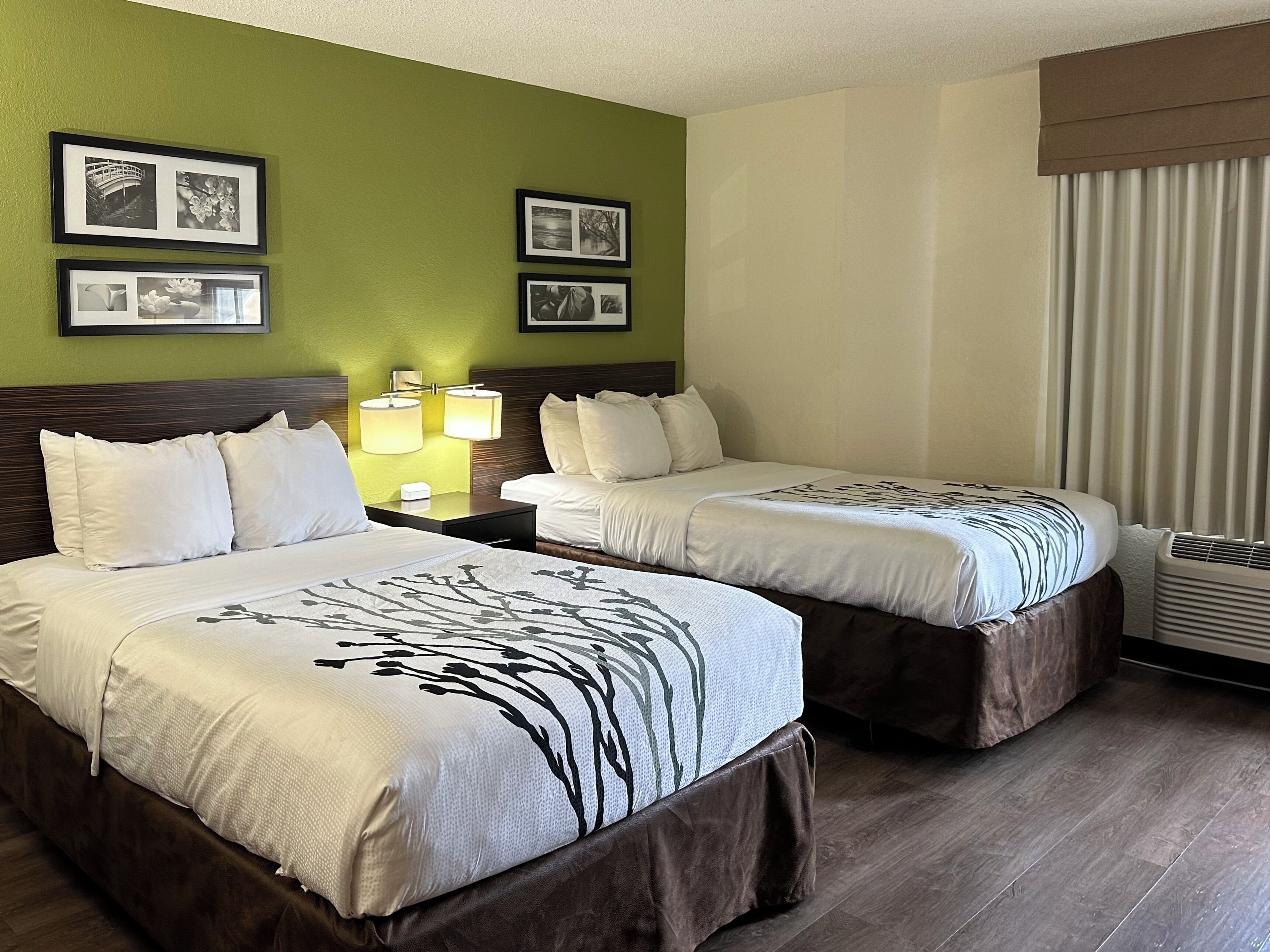Une chambre d’hôtel avec deux lits, une table de chevet avec des lampes, des tableaux encadrés au mur et un appareil de climatisation.