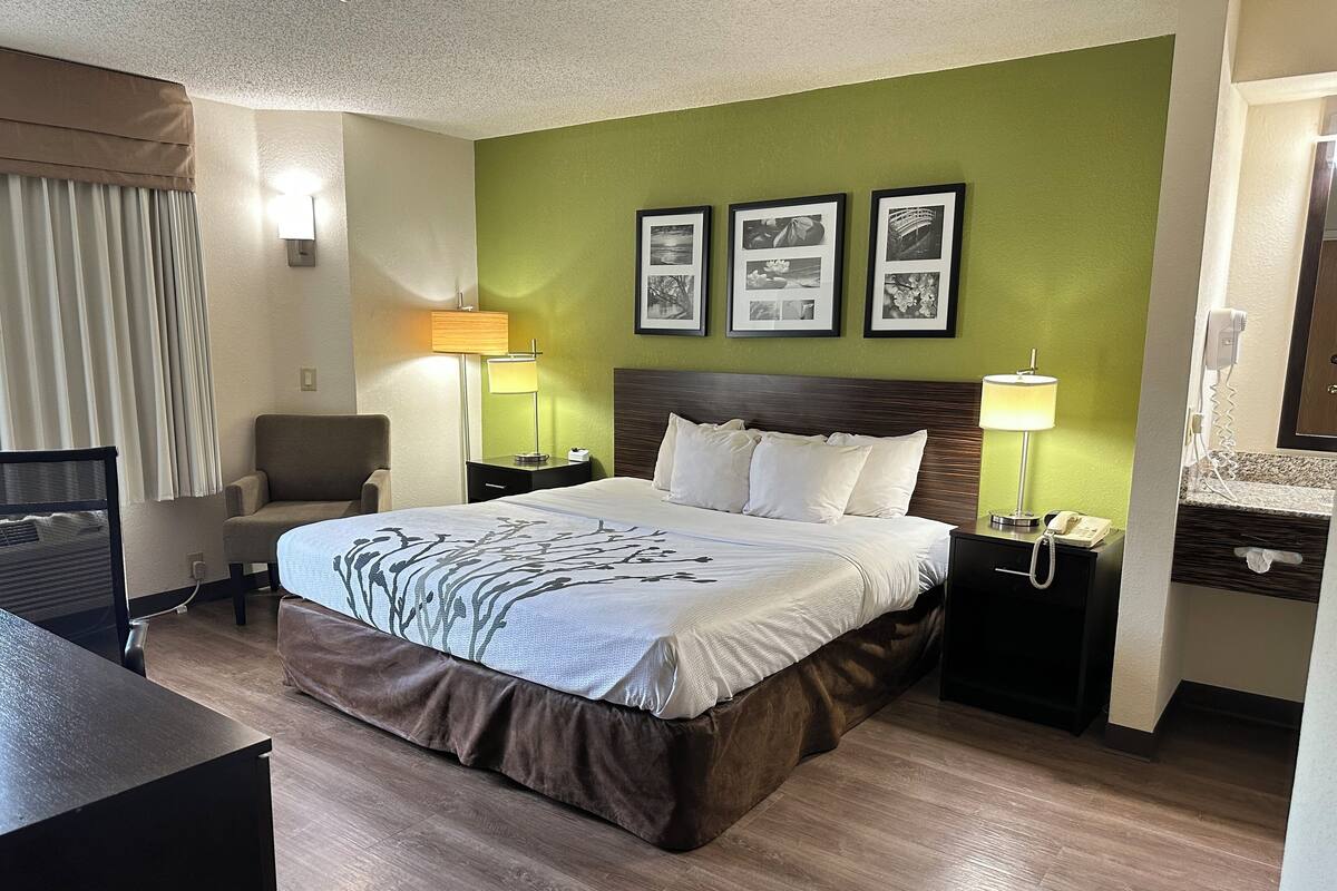 A hotel room with a large bed, a desk, a chair, a nightstand with a lamp, and a wall with framed pictures.