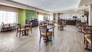 Un amplio comedor de hotel con varias mesas y sillas, una cocina al fondo y un gran ventanal que proporciona luz natural.