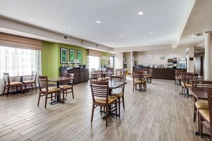 Un amplio comedor de hotel con varias mesas y sillas, una cocina al fondo y un gran ventanal que proporciona luz natural.