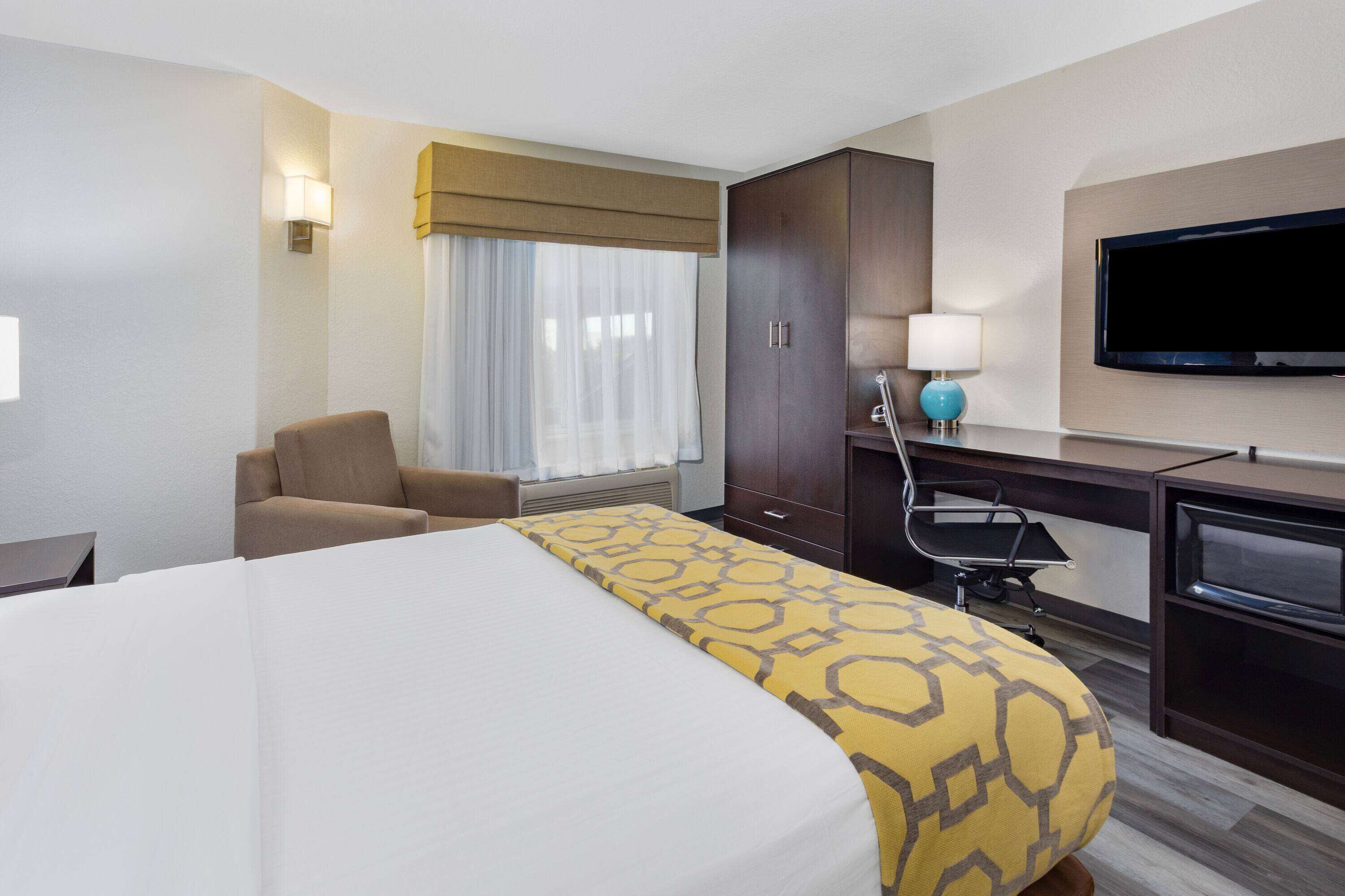 A hotel room with a large bed, a desk with a computer, a chair, a TV, and a window with curtains.