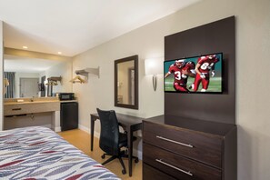 Quarto de hotel com cama, escrivaninha, cadeira, televisão, espelho e um pequeno frigorífico.