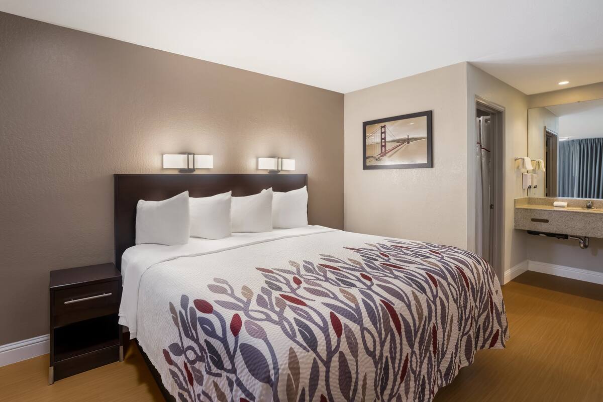 A hotel room with a large bed, two bedside lamps, a nightstand, a bathroom with a sink and mirror, and a framed picture on the wall.