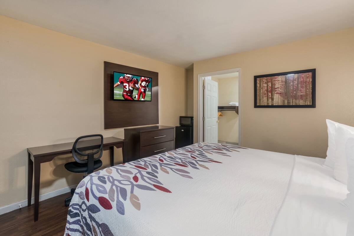 A hotel room with a bed, a desk, a chair, a TV, and a painting on the wall.