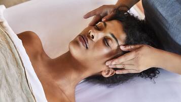 A person lying on a bed with a white pillow and a white sheet, receiving a head massage.