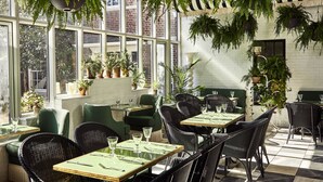 A restaurant with green upholstered chairs and tables, large windows, and hanging plants. - The Vanderbilt, Auberge Collection (Newport)