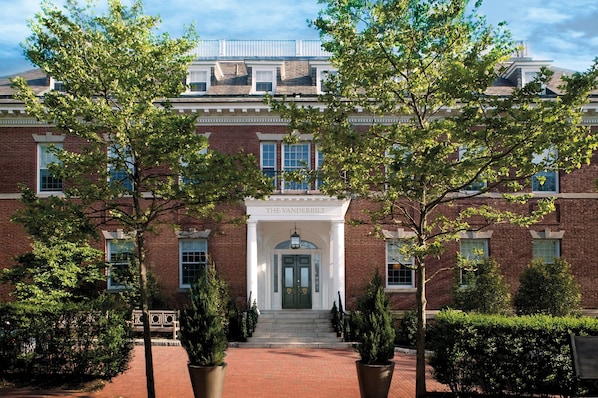 A brick building with a central entrance, surrounded by trees and a pathway leading to the entrance.