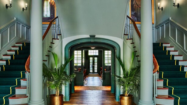 A grand hallway with a wooden floor, flanked by two staircases with red and white steps, leading to a door at the end. - The Vanderbilt, Auberge Collection (Newport)