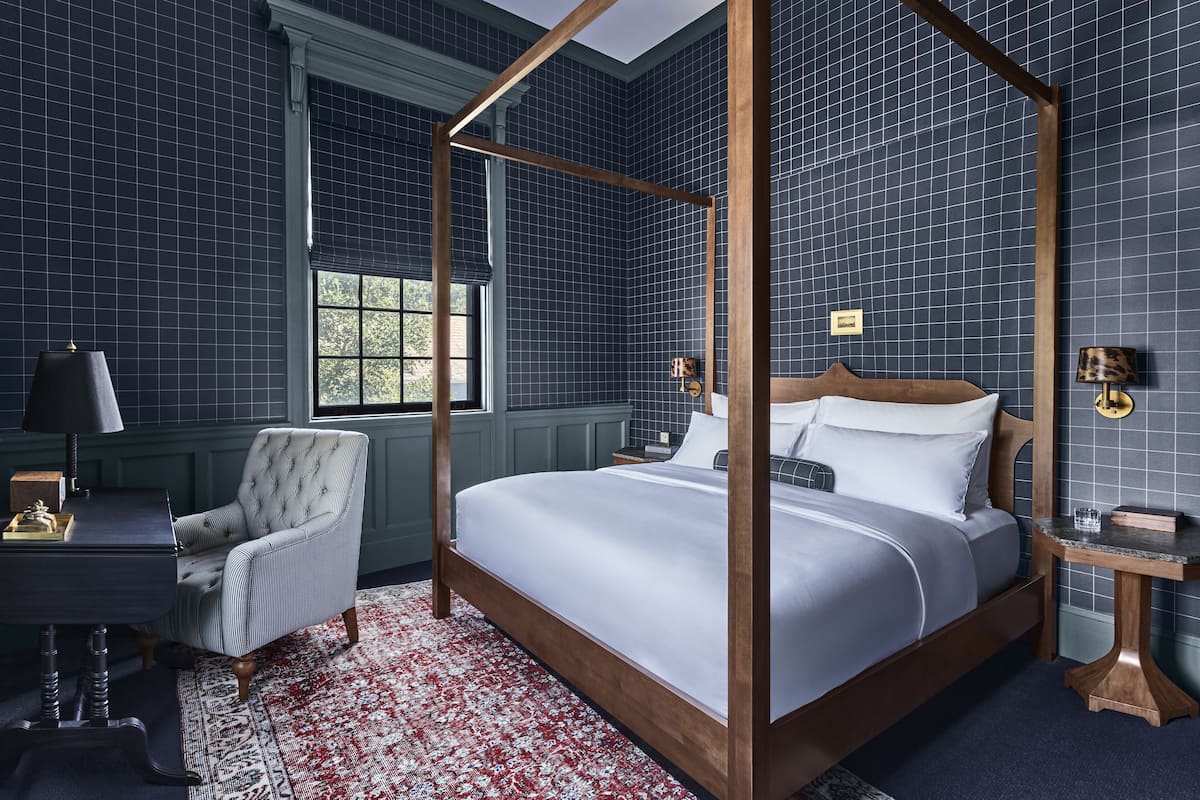 A four-poster canopy bed with a wooden frame and white linens, a tufted armchair, a desk with a lamp, and a patterned rug.
