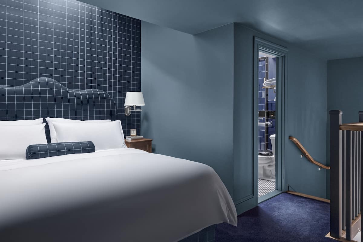 A hotel room with a large bed, a nightstand with a lamp, a view of the bathroom through a door, and a wooden staircase.