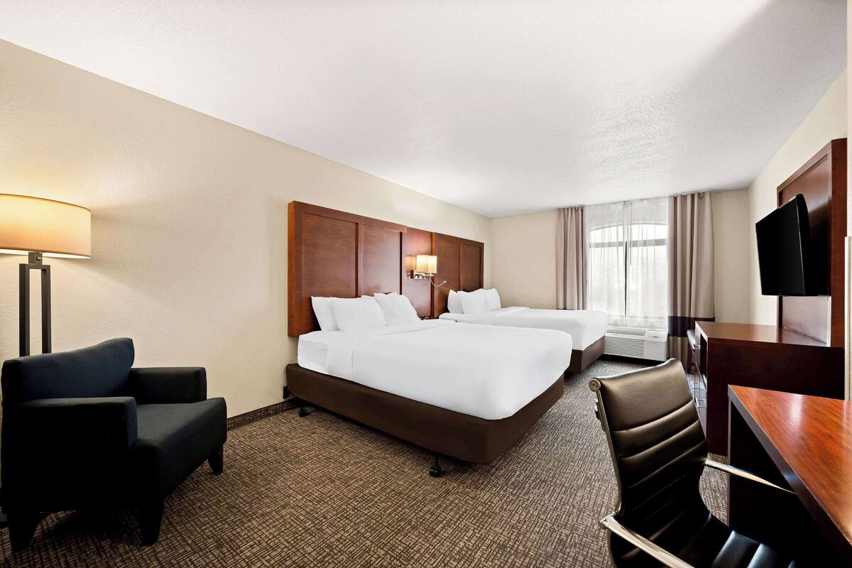 A hotel room with two beds, a desk, a chair, a TV, and a lamp.