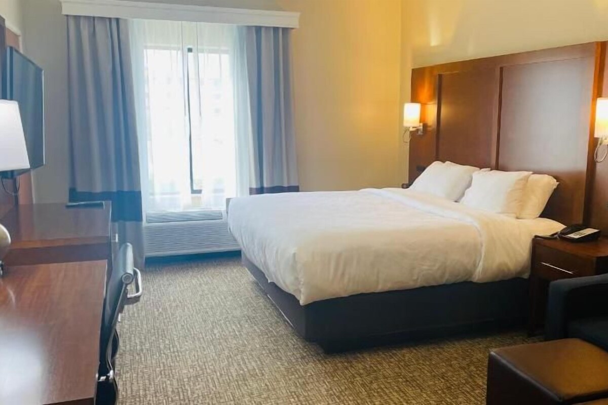 Ein Hotelzimmer mit einem Bett, Nachttischen, einem Schreibtisch mit Lampe, einem Sessel und einem großen Fenster mit Vorhängen.