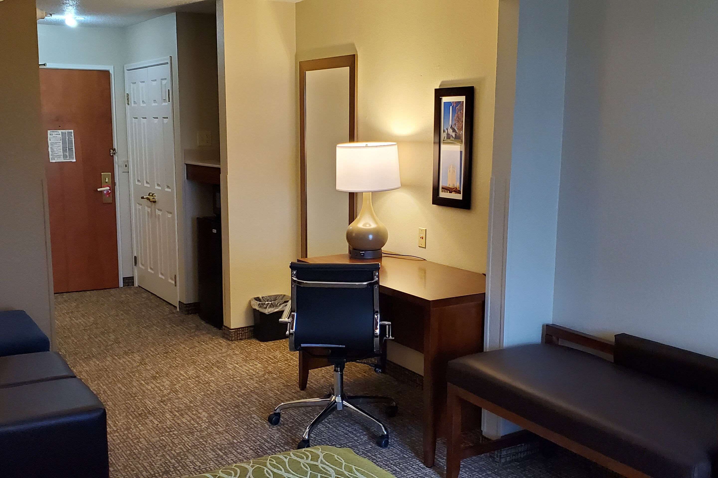 Una habitación de hotel con escritorio, silla, lámpara, banco y cubo de basura.
