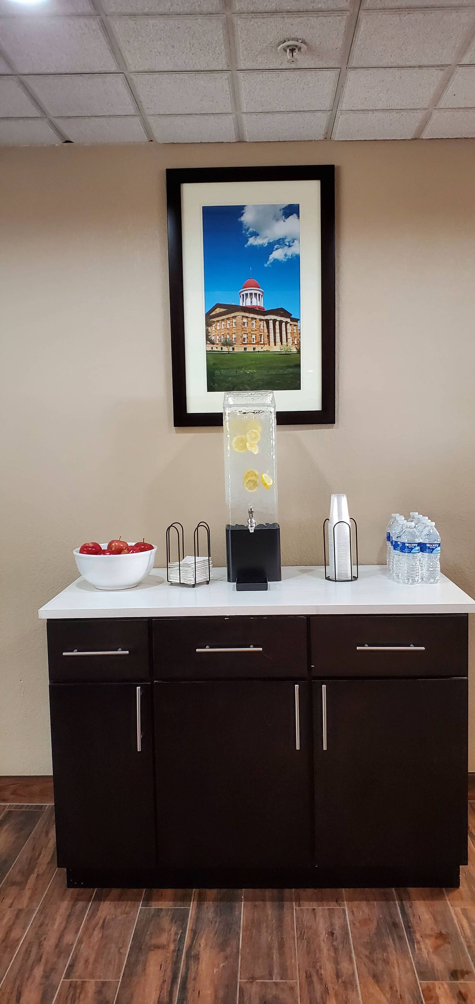 A reception desk with a framed picture of a building, a water dispenser, a bowl of apples, and a stack of magazines.