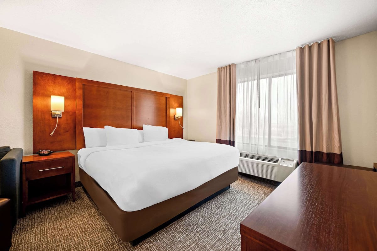 A hotel room with a large bed, a wooden headboard, two bedside tables with lamps, a desk, and a window with curtains.