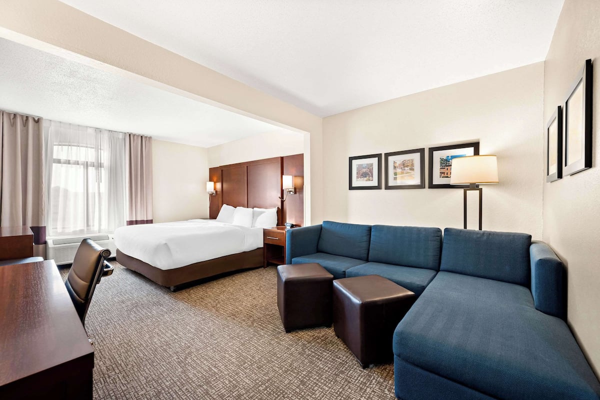 A hotel room with a large bed, a sofa, a desk, and a chair. There are framed pictures on the wall and a lamp on the desk.